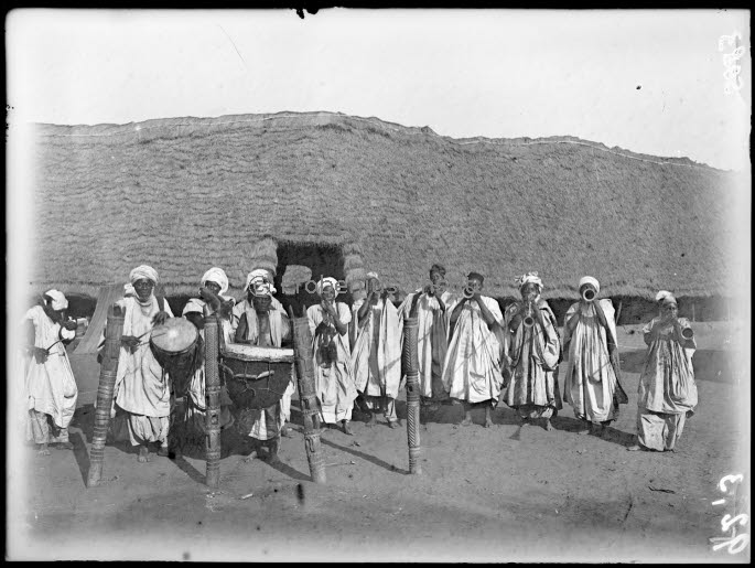 A group of people dressed in traditional robes and head coverings stand in front of a large, dome-shaped thatched structure with a small central entrance. Some individuals hold musical instruments, including drums and long wind instruments, suggesting a cultural or ceremonial performance. The scene is set outdoors on bare earth, evoking a traditional village atmosphere.