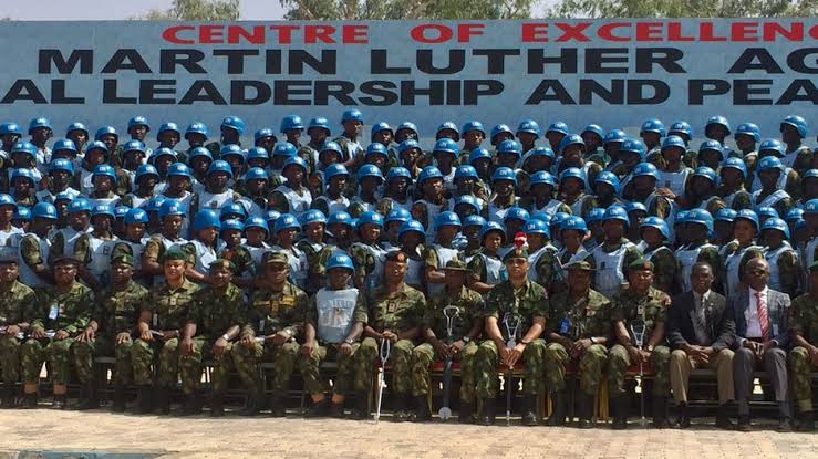 A large group of military personnel in blue helmets pose for a group photo in front of a sign that reads “CENTRE OF EXCELLENCE MARTIN LUTHER AG NATIONAL LEADERSHIP AND PEACE.” Some individuals are seated while others stand behind them, suggesting a formal event or graduation ceremony.
