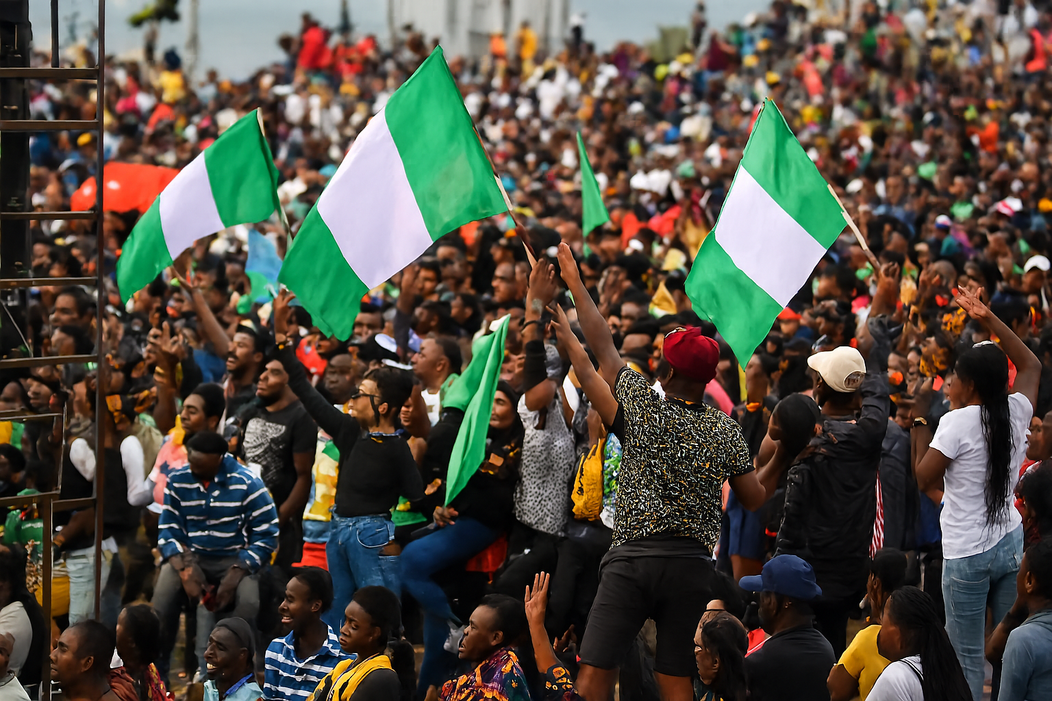 A large crowd of Nigerians gathers outdoors during the day, waving multiple green-white-green Nigerian flags. The people, diverse in age and attire, raise their hands and flags in a show of unity and activism. In the foreground, a young man in a red beret and leopard-print shirt holds a flag aloft, while others around him cheer and gesture. The background reveals more participants, a speaker tower, and a hazy shoreline in the distance. The image is vibrant, with enhanced clarity and rich colors highlighting the energy and solidarity of the scene.