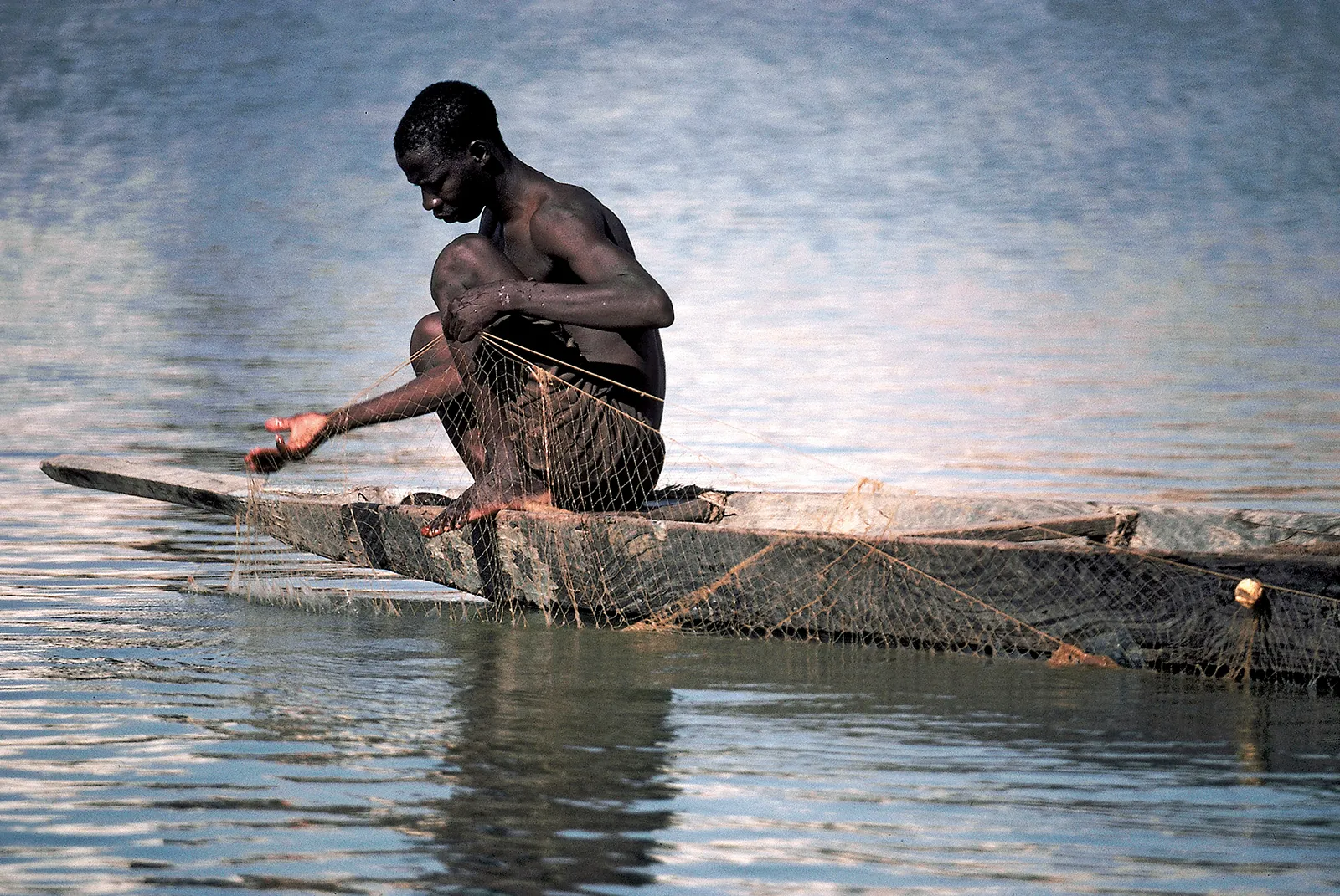 A person crouches on a narrow wooden boat floating on calm, reflective water, carefully handling a fishing net that drapes over the side and dips into the water. The scene captures a moment of focused, traditional fishing activity in a tranquil setting.