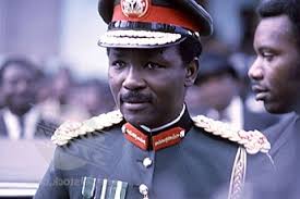 "High-ranking military official in formal uniform with gold epaulettes, red collar insignia, and medals, wearing a peaked cap with a gold emblem, standing in a public setting during a ceremonial event.
