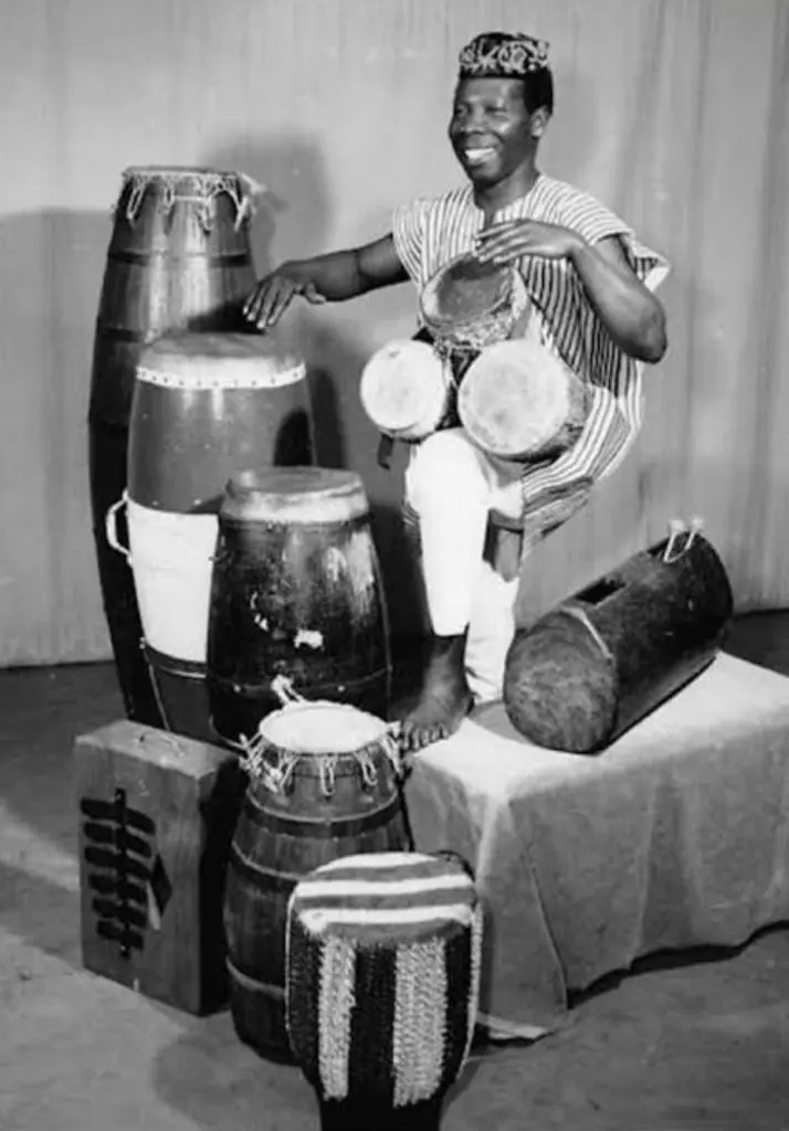 An individual dressed in a striped traditional garment and patterned hat sits surrounded by various traditional African drums and percussion instruments. They are actively playing two small hand drums positioned in front of them. The setup includes cylindrical and barrel-shaped drums, as well as a rectangular wooden slit drum, all arranged on a woven mat. The scene highlights a cultural demonstration of African musical heritage.