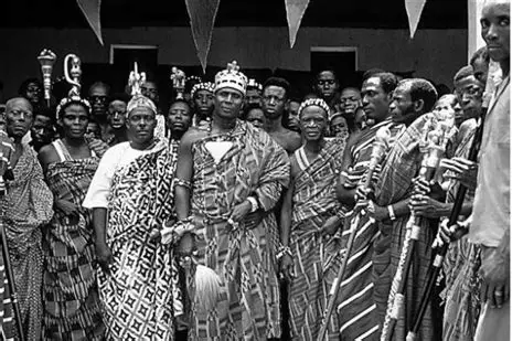 A vibrant cultural gathering featuring individuals dressed in traditional, patterned attire. Some participants hold ceremonial staffs or objects, suggesting a ritual or celebratory event. Triangular flags are strung overhead, adding to the festive atmosphere. The scene highlights cultural heritage and communal participation.