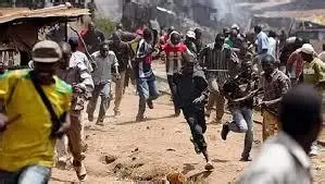 A chaotic outdoor scene with people running in various directions amid makeshift structures and scattered debris. Smoke rises in the background, suggesting a disturbance or emergency. The individuals wear casual clothing, and some carry objects, conveying urgency and tension.