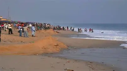 Crowded beach scene with people standing on the sand and in the water, under a clear sky with gentle ocean waves and visible shoreline erosion.