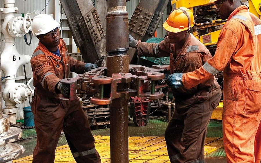 Three industrial workers operate heavy machinery at an oil drilling site. Dressed in protective gear including helmets and orange or brown coveralls, they work together to manipulate a large vertical pipe using a specialized tool. The environment is filled with complex equipment, piping, and metal structures, emphasizing the technical and physically demanding nature of oil rig operations. The scene captures teamwork, precision, and safety in a high-risk industrial setting.