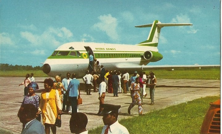A group of passengers, including uniformed personnel and individuals carrying luggage, board or disembark from a white Nigeria Airways aircraft with green stripes on an airport tarmac. The fuselage displays the airline's name, and the background features a partly cloudy blue sky and grassy terrain.