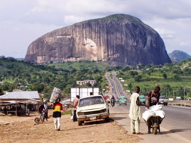 A massive granite monolith dominates the landscape, rising dramatically from green forested hills under a cloudy sky. In the foreground, a paved road runs toward the rock formation, with several people visible along the roadside - some standing and others bent over, appearing to be engaged in various activities. Vehicles including cars and what appears to be a van are parked along the road. Simple buildings and structures are scattered in the middle distance between the road and the imposing rock face. The scene appears to be in a rural or semi-rural African setting, with the monolith serving as a prominent natural landmark.