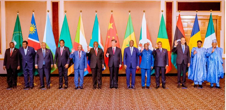 Fourteen international dignitaries stand in a straight line, each positioned in front of a national flag representing countries such as China, Namibia, and the African Union. They are dressed in formal attire, with some raising their hands or waving, suggesting a moment of greeting or celebration. The setting is a formal venue, likely for a diplomatic or international event focused on global cooperation.