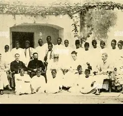 A vintage group photograph of about 26 people, mostly dressed in white traditional attire, posed outdoors in front of a building with an archway and hanging greenery. Some individuals are seated while others stand behind them.