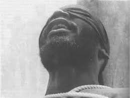 Black and white image of Baba Oni Lace, a bearded man wearing a blindfold and head covering, with his head tilted back and mouth slightly open; a rope is visible around his neck, conveying a vulnerable and restrained state.