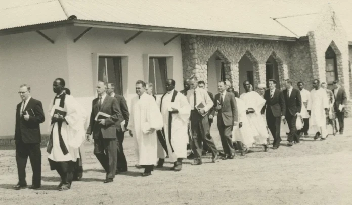 A black and white photo of a religious procession outside a stone building with arched windows, likely a church. The group includes individuals in religious robes and formal suits, walking solemnly while carrying books, possibly hymnals or religious texts. The image evokes a sense of tradition and ceremony.