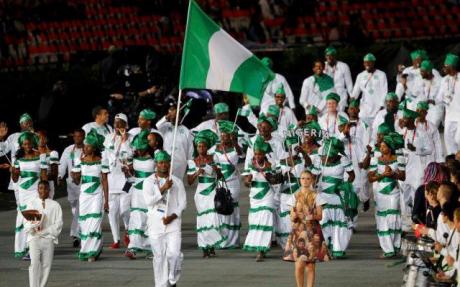 A group of people dressed in coordinated white outfits with green accents march in formation during a parade, holding a large Nigerian flag. They wear matching headgear, and one person leads as the flag bearer. The setting suggests an international event, possibly an Olympic opening ceremony, highlighting national pride and unity.