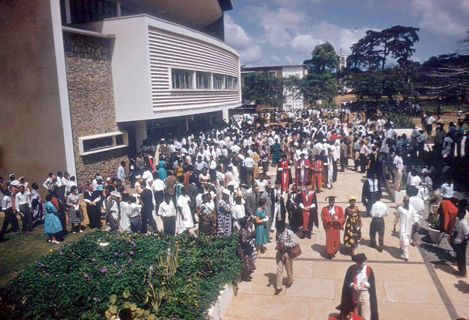 A large crowd gathered outside a modern building with stone and white walls, many dressed in academic gowns and caps, indicating a graduation ceremony. The scene includes people in traditional and modern attire, landscaped greenery and flowers in the foreground, and a partly cloudy sky above.