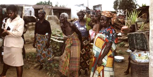 A group of people gathered outdoors in a rural setting, wearing a mix of traditional and modern clothing. Colorful patterned fabrics are visible among the attire. The background features wooden structures, vegetation, and household items like bowls and a chair, suggesting a community gathering or event.